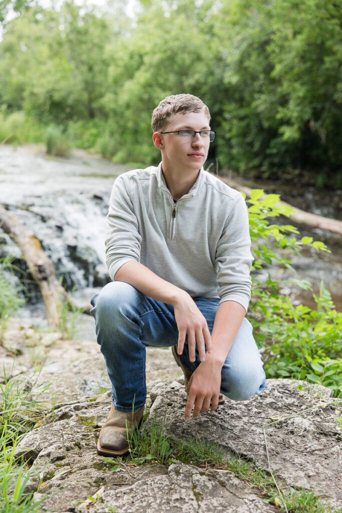 senior guy kneeling by a waterfall looking off into the distance