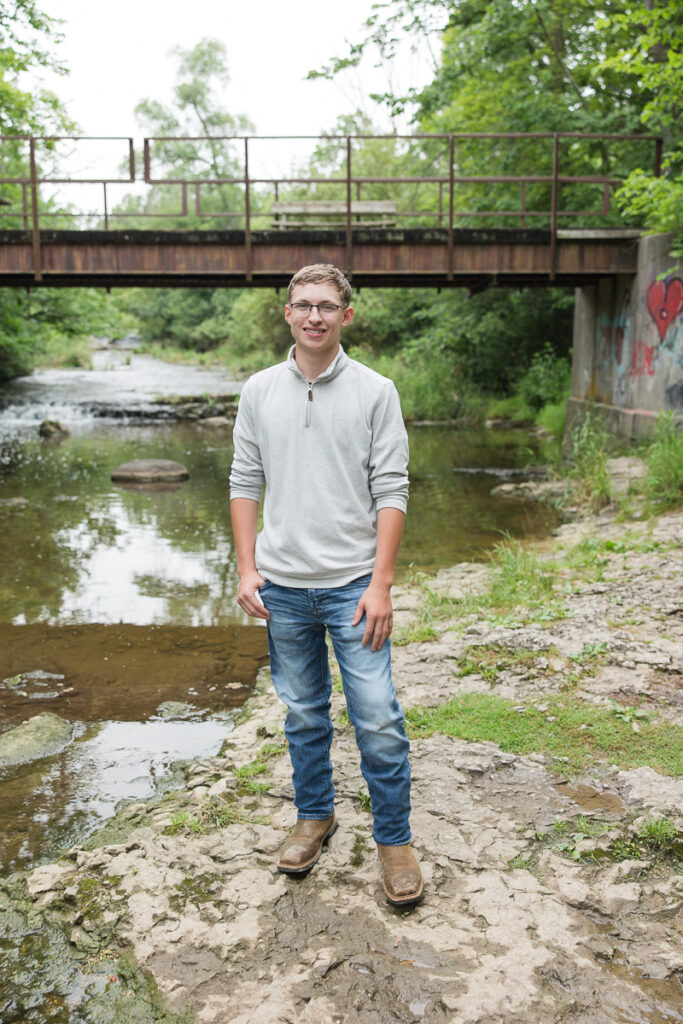 senior guy standing in front of a bridge along a creek smiling at the camera